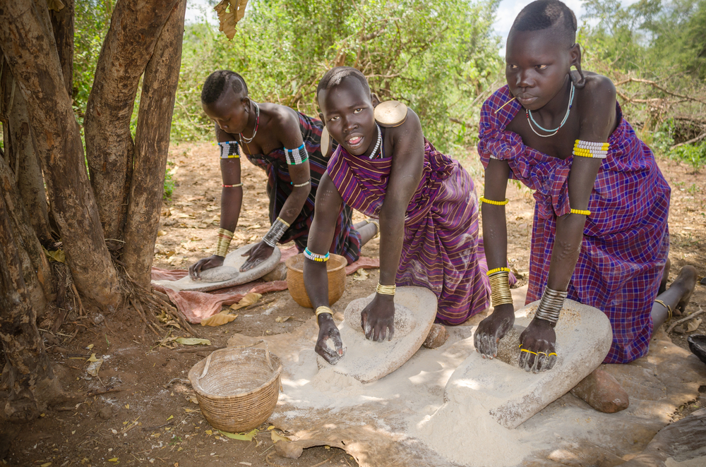 girls from Mursi tribe grinding cereals with a stone to prepare flour