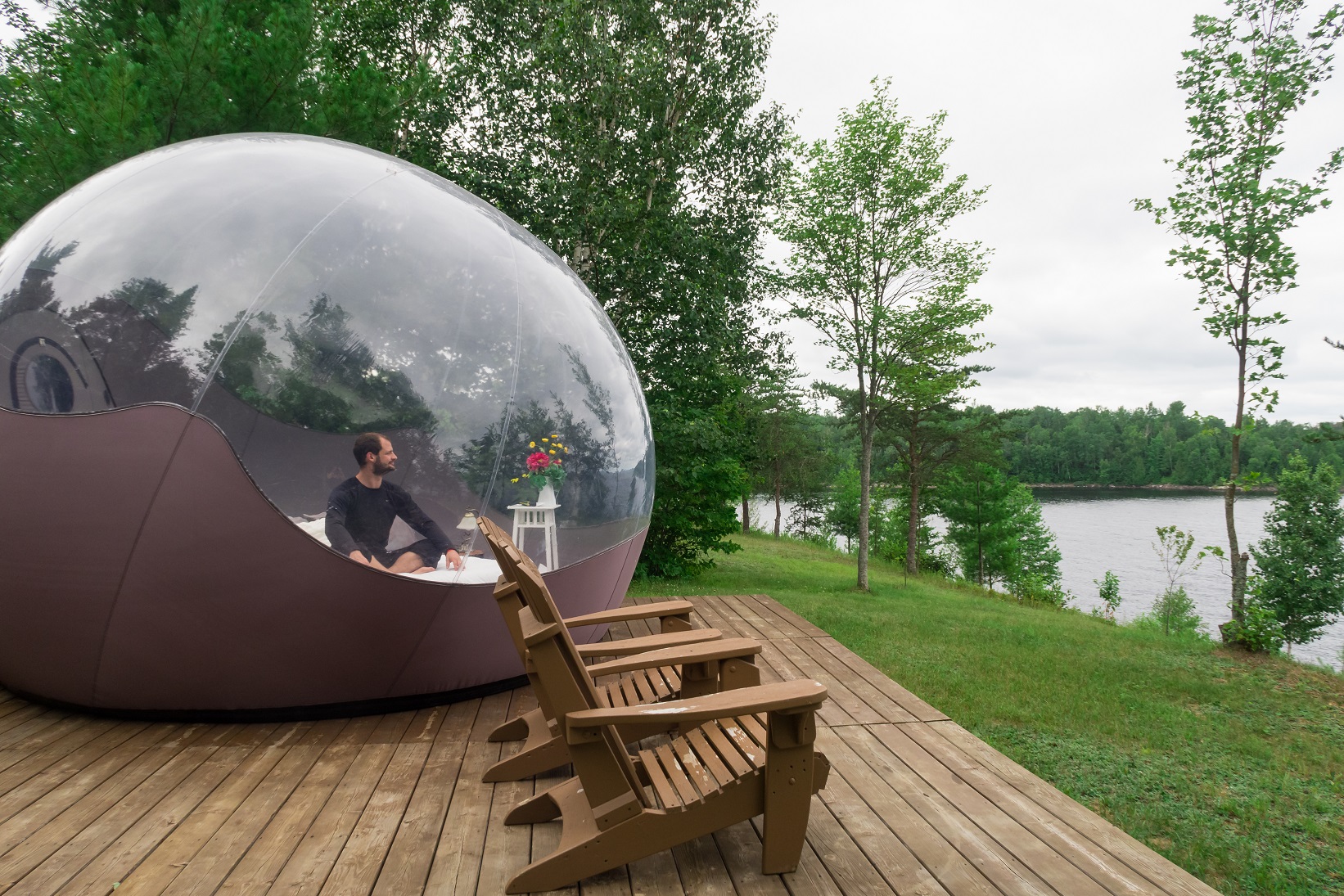 Man sitting on a bed inside a transparent bubble tent facing a lake - 2020