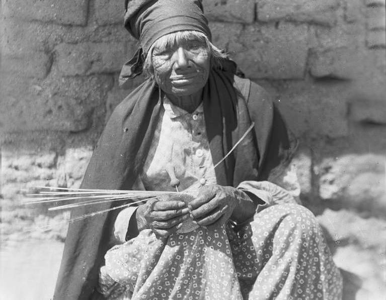 Cahuilla woman sits in front of brick wall with a partially completed basket in her hands