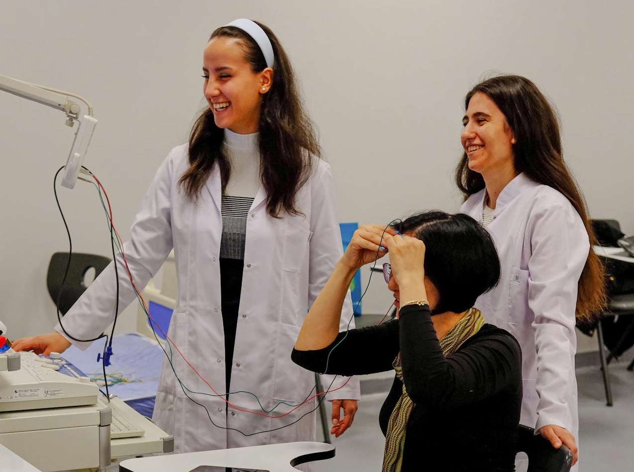 Two Women in Lab Coats and a Woman Sitting on a Chair in an Examination Room