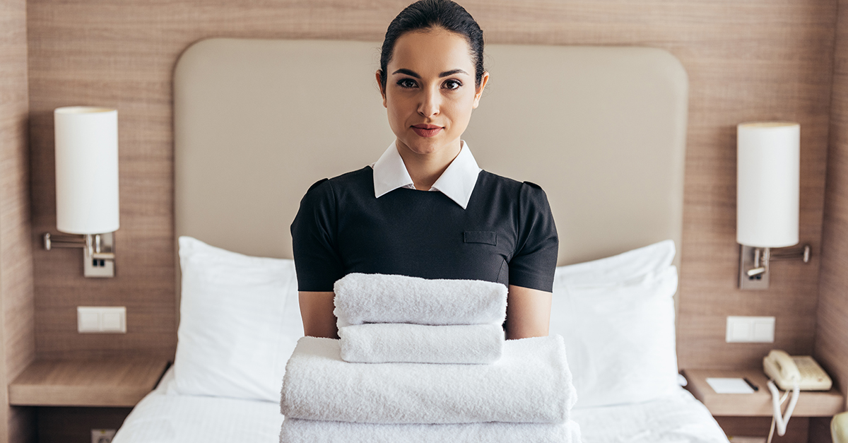 front view of maid holding pile of folded towels near bed and looking at camera in hotel room