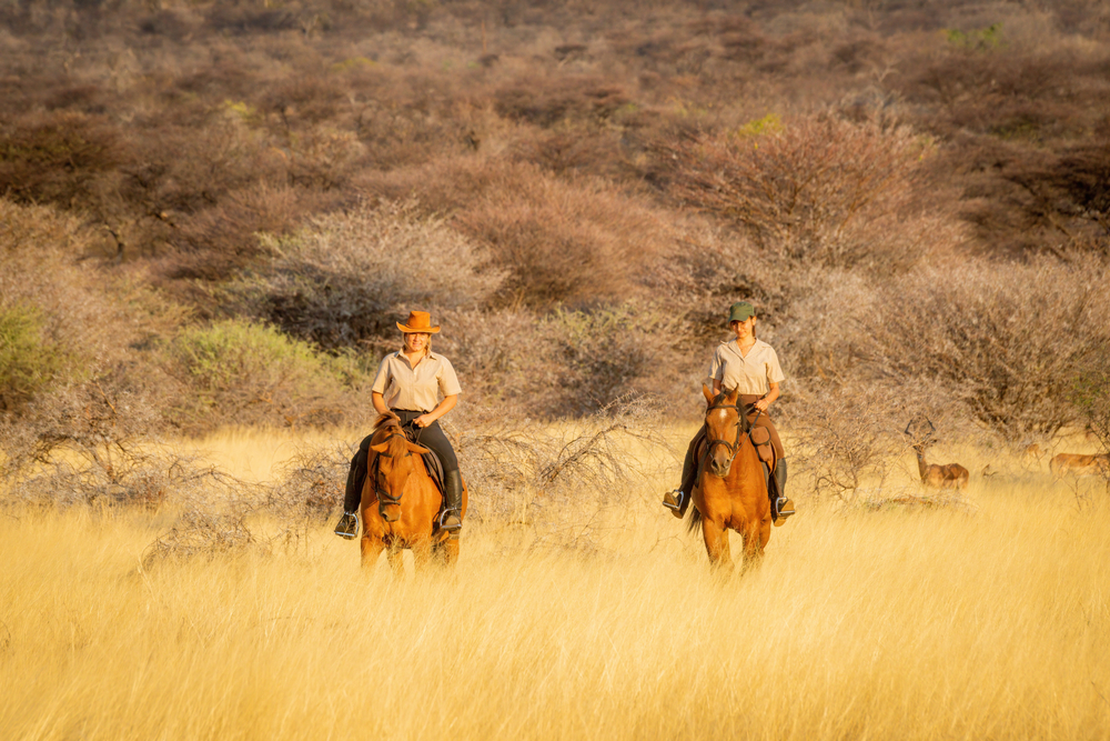 Two women ride horses watched by impala