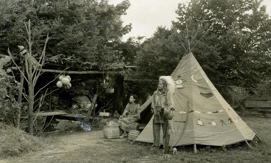 Grayscale photo view of two Cherokee in traditional dress outside of a teepee
