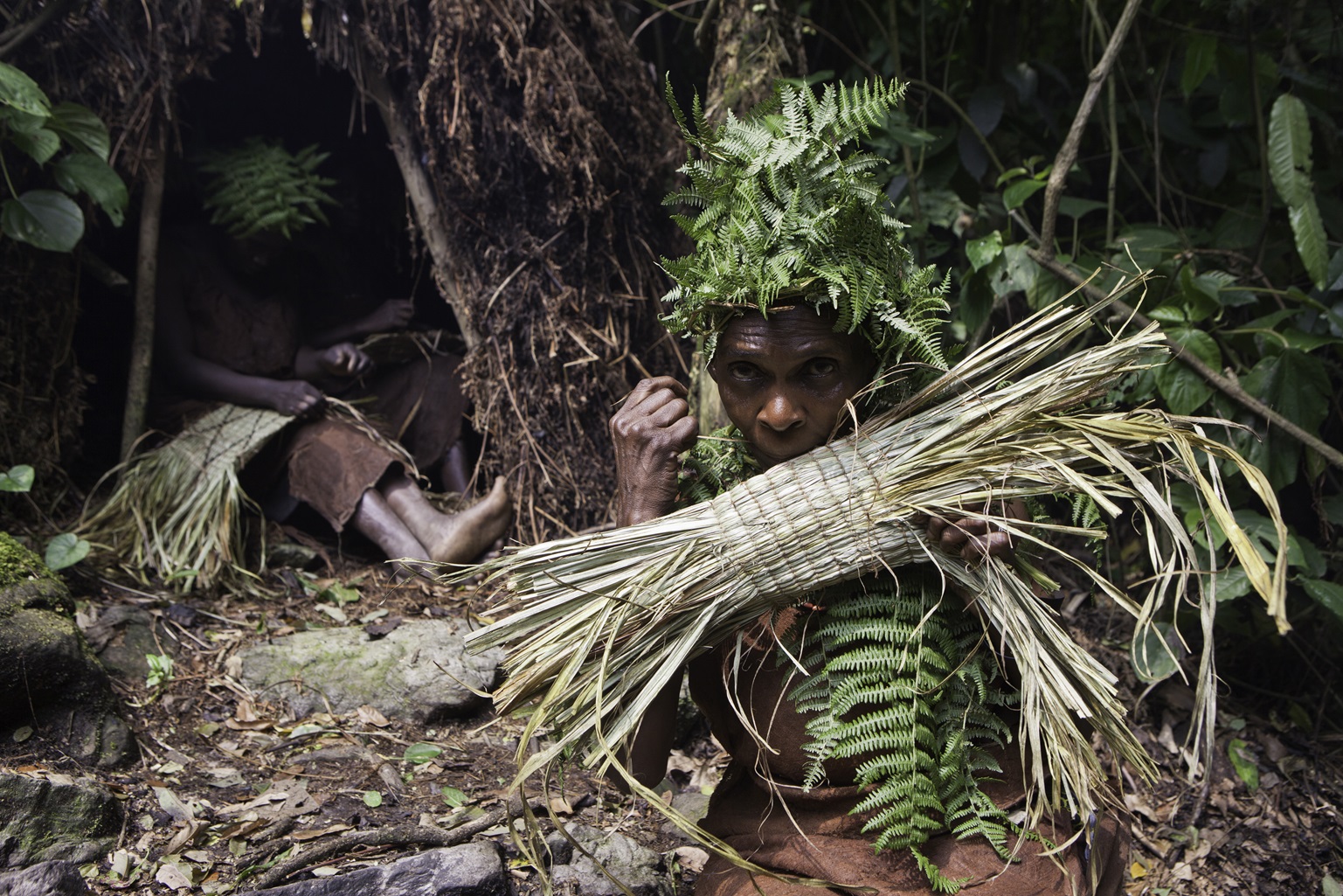 A Batwa woman in traditional dress uses reeds to make a basket.