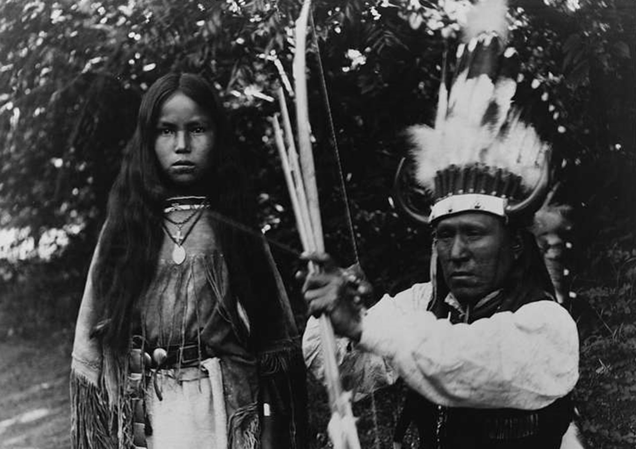 Elk Tongue, wearing war bonnet while crouching with bow and arrow in hands - 1891
