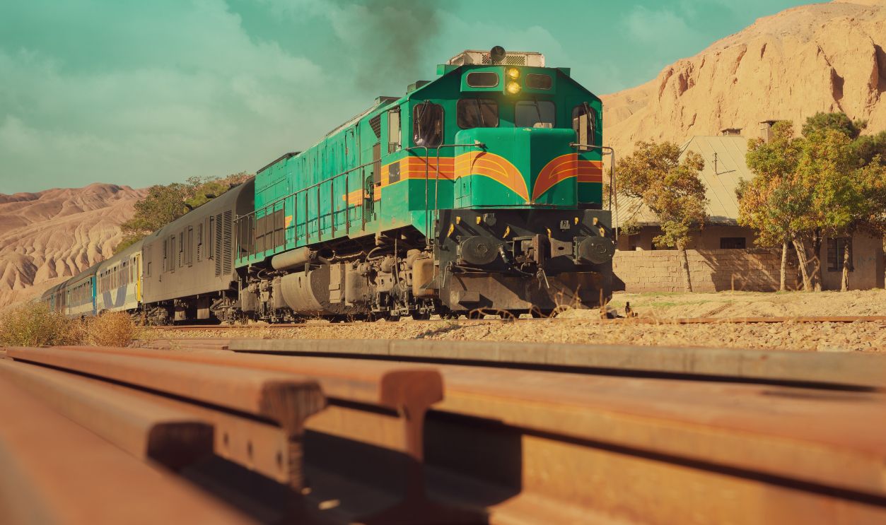 An amazing view of a green GM diesel locomotive traveling on the Trans-Iranian railway. There are a bunch of worn-out rails in the foreground of the frame.