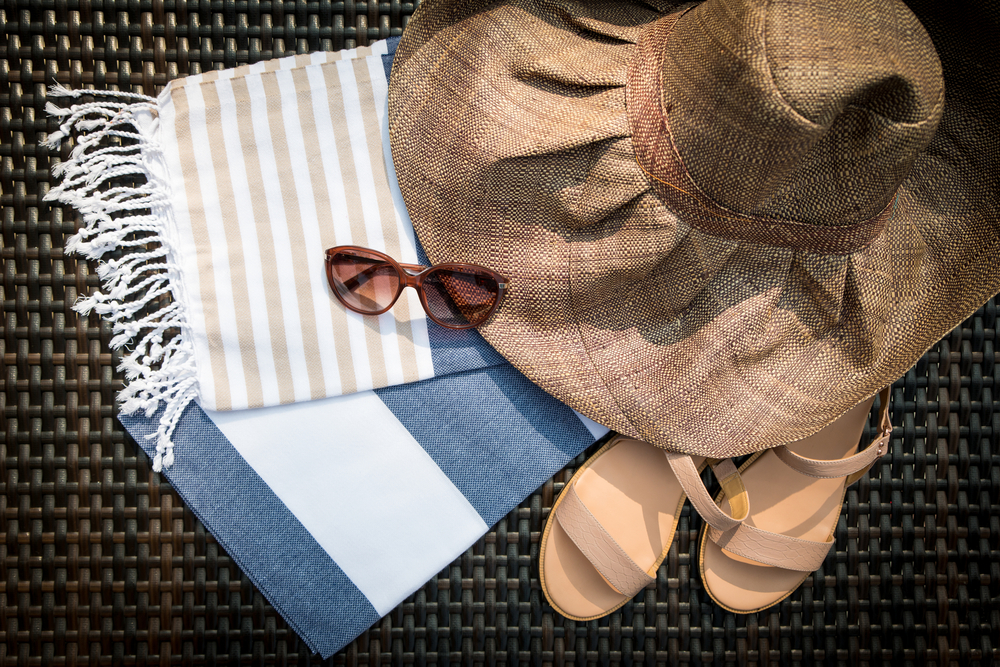 Summer accessories of white, blue and beige Turkish towel, sunglasses, beige leather ladies sandals and straw hat on a rattan lounger.