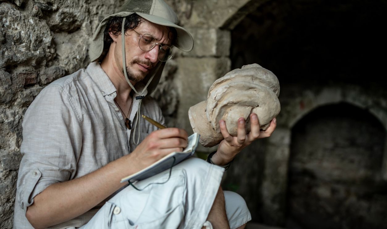 Gettyimages - 2218936978, Archaeologist Studying Ancient Artifact in a Historical Site - stock photo Focused archaeologist examining and sketching an artifact in a historical setting, showcasing passion and precision in historical studies and preservation.