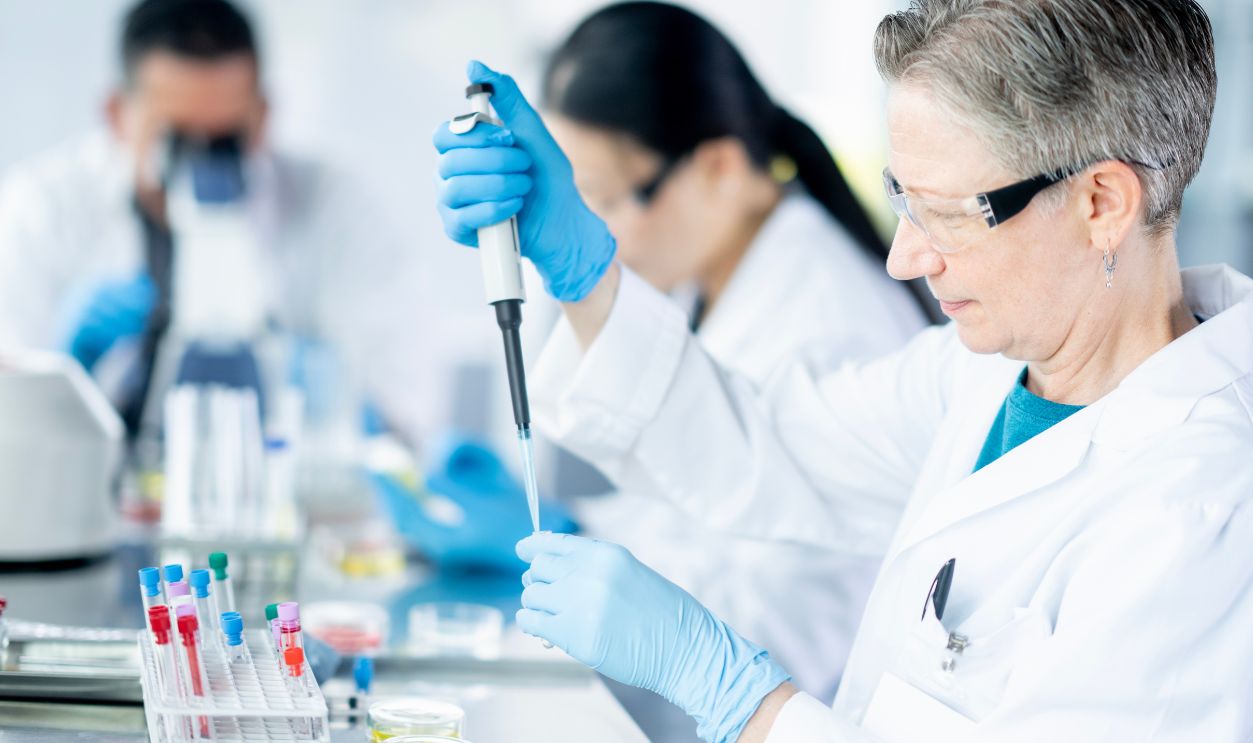 Gettyimages - 2181815202, Analyzing the Data - stock photo A small team of adults work together in a Medical Lab as they draw samples and take a closer look under microscopes. They are each dressed professionally in lab coats and are wearing protective eyewear as they analyze the data.