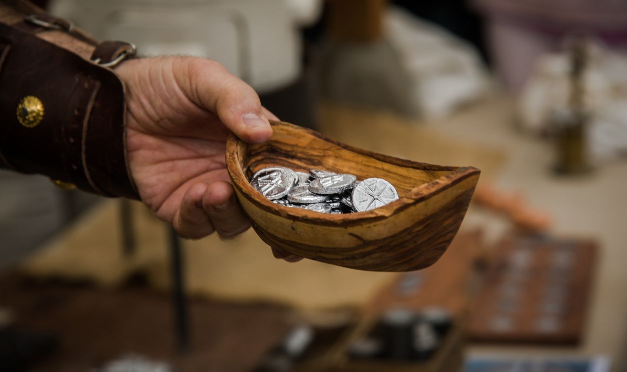 A person in leather armor holds a bowl filled with Roman coins, with a coin-making workshop visible in the background.
