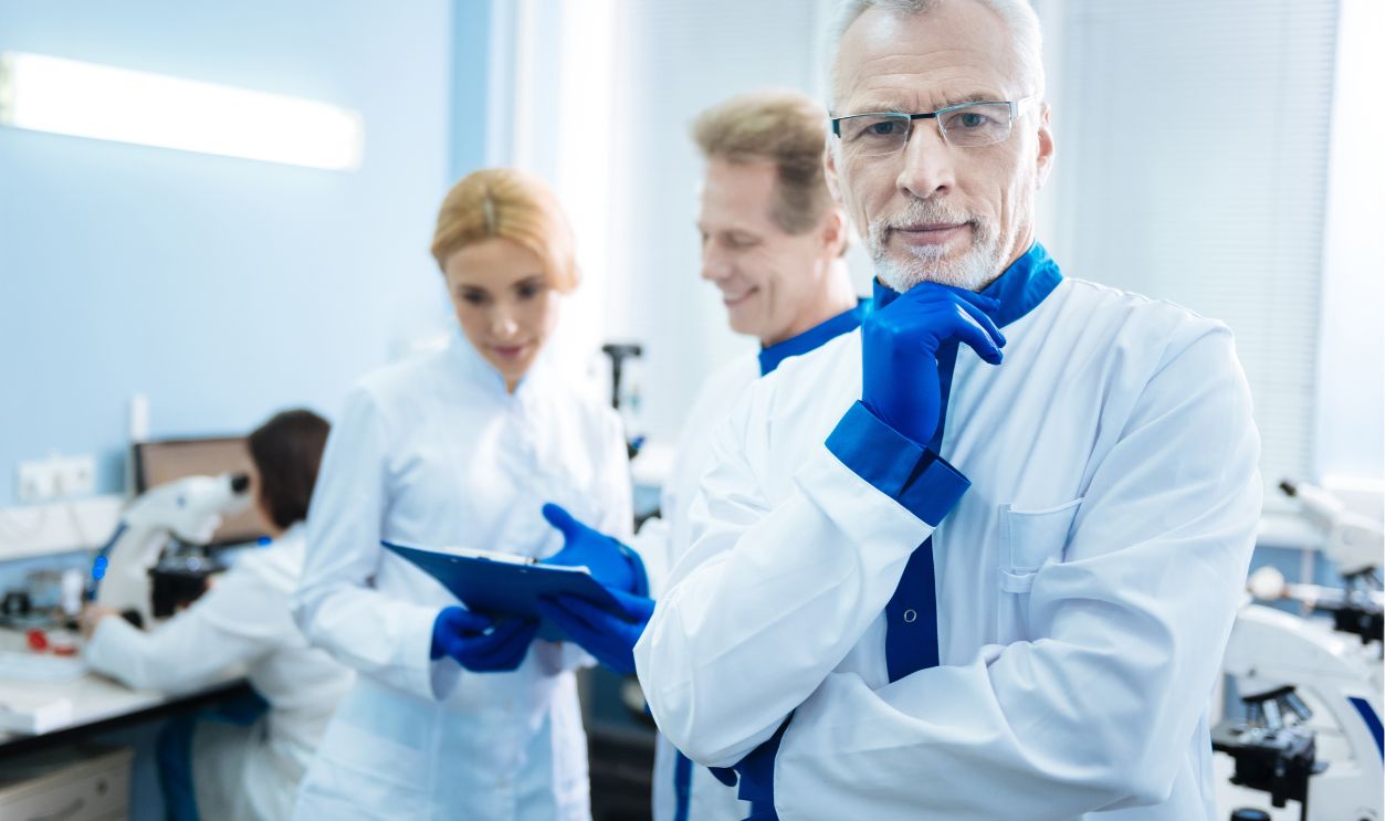 Working atmosphere. Nice grey-haired wrinkled researcher wearing a uniform and touching his chin while others discussing something in the background