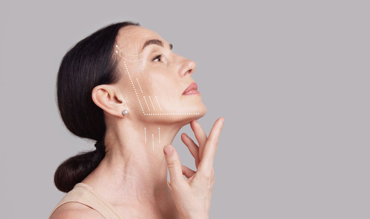 Beautiful senior woman with lifting arrows showing facial anti-aging treatment on skin against grey studio background. Negative space. Concept of beauty procedures, cosmetology treatment, massage. Ad