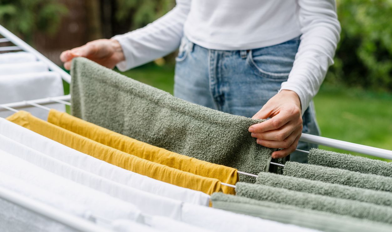 cropped shot of female hands hang fresh clothes on metal drying rack in backyard after laundry or housewife in blue jeans doing housework routine