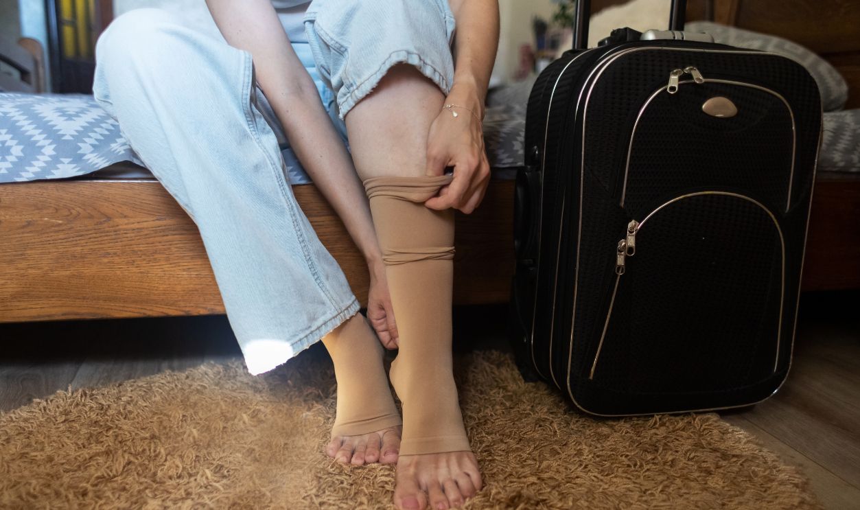 Woman putting on compression stockings before a long trip, suitcase in the background. Concept of leg protection during travel, varicose veins and deep vein thrombosis on long flights or car journeys