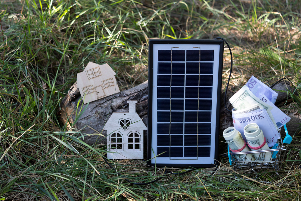 There is a solar panel on the grass, two mock-up houses and money in a supermarket trolley.