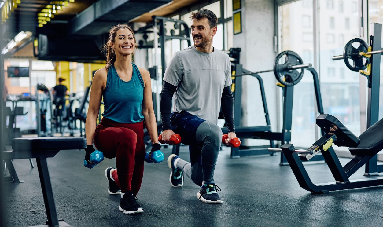 Happy athletic couple exercising with hand weights in lunge position in a gym