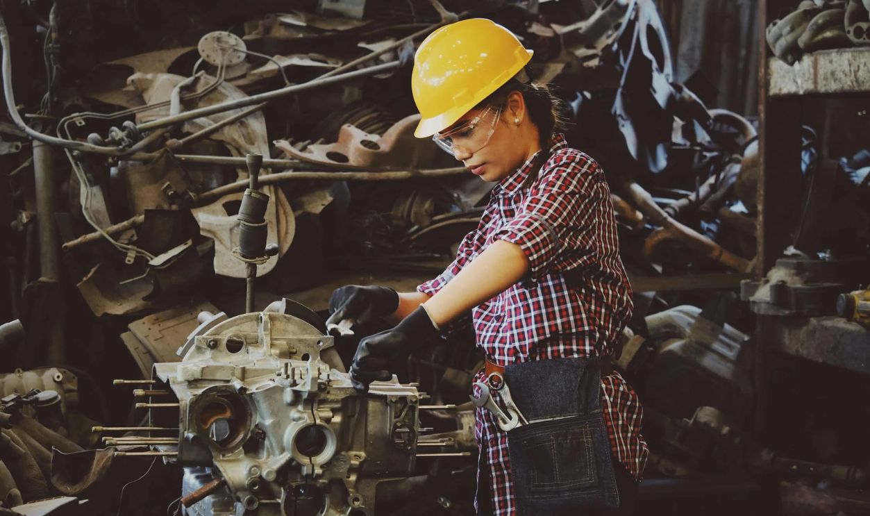 Woman Wears Yellow Hard Hat Holding Vehicle Part