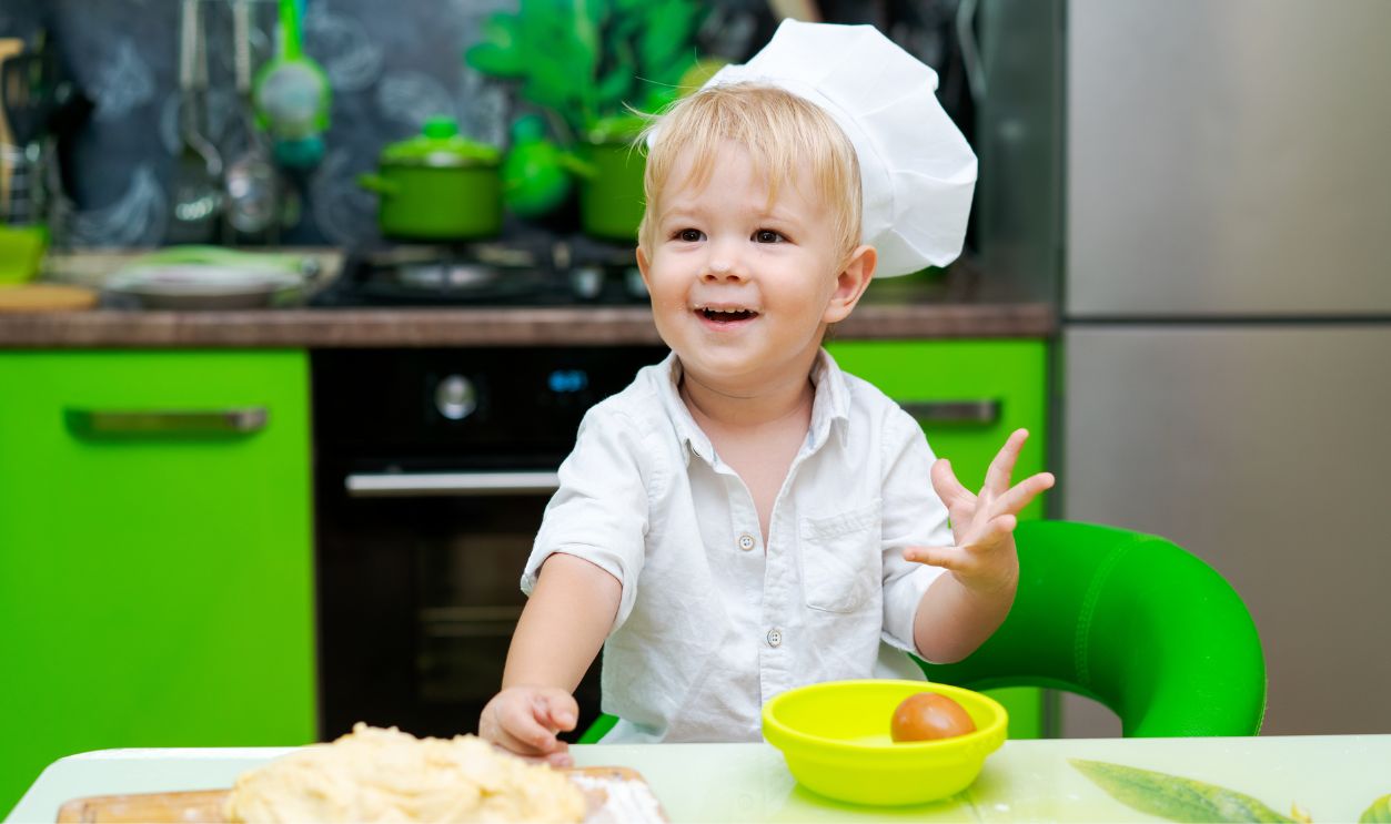 happy little boy preparing dough in kitchen at table. there are dough products on table, dressed as chef