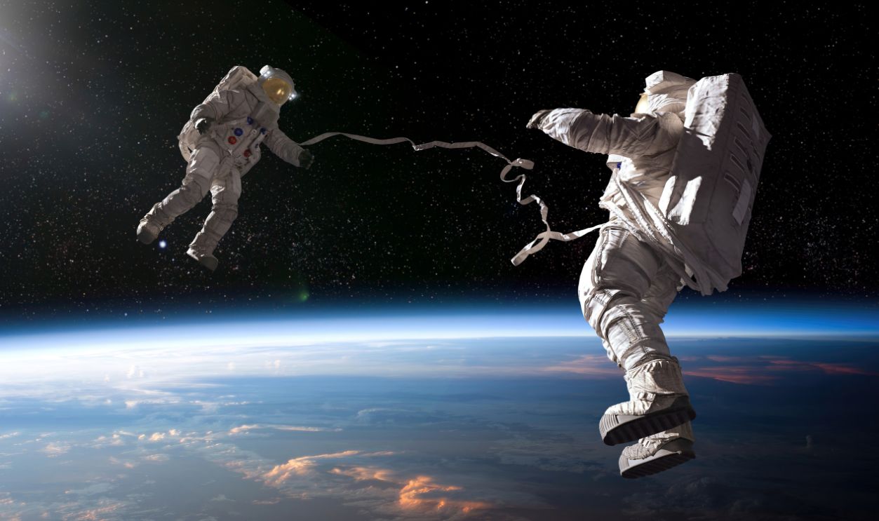 Two astronauts in full spacesuits with backpacks on a space walk with tethers, facing each other with hands out. The astronauts are in front of planet earth and some distant stars are also visible. Earth image from NASA