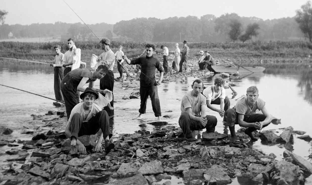 Gettyimages - 526305138, Young men fishing in a stream, ca. 1915. A group of men line a rocky stream and fish with cane poles.