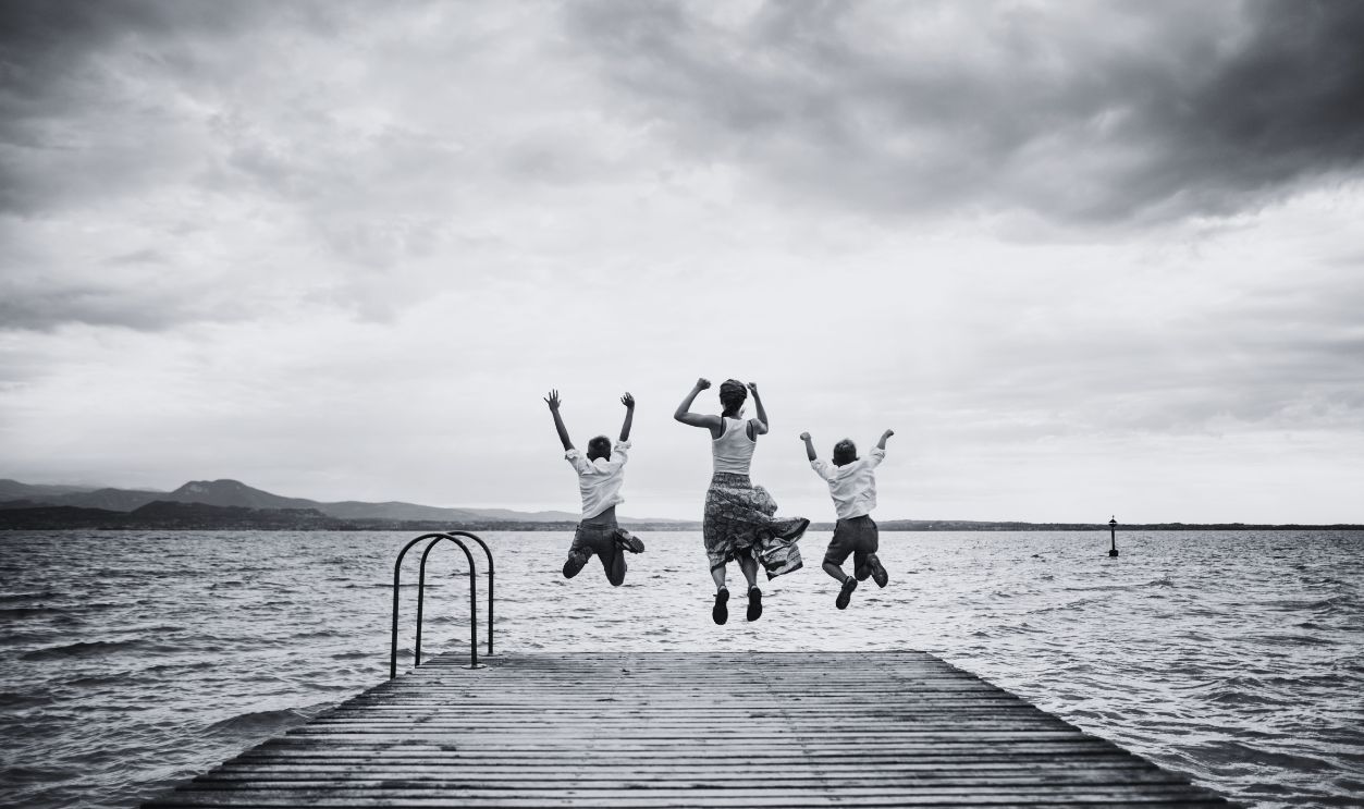 Three kids jumping off a wooden dock into a calm lake - stock photo Kids enjoying Garda Lake vacations. Three children mid-air as they leap from a sturdy wooden dock into a tranquil body of water on an overcast summer day.