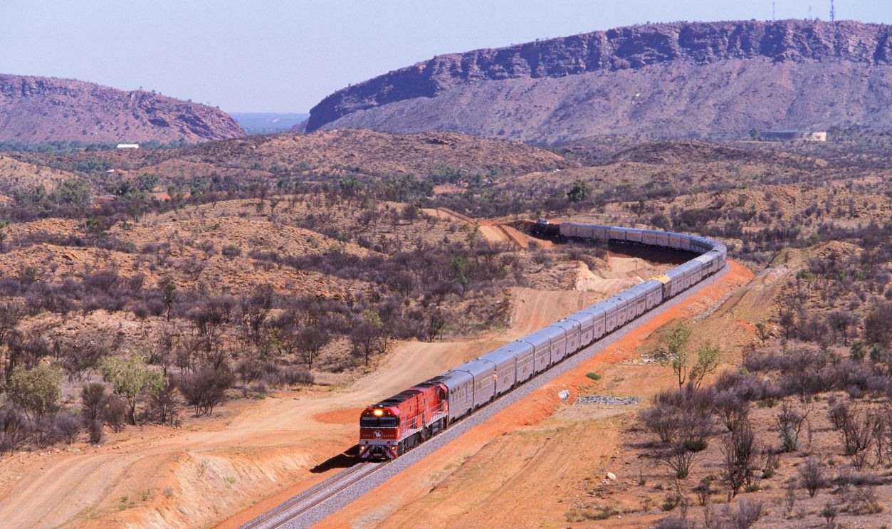 "Alice Springs, Australia - February 2, 2004: The first Ghan passenger train to Darwin departs Alice Springs with the spectacular MacDonnell Ranges in the background. The extra long VIP train is mid-way through its inaugural 2969km transcontinental journey from Adelaide in the South to Darwin in the North. The first train departed Adelaide February 1 and arrived in Darwin to a huge welcome on February 3, 2004. The journey from the Mediterranean South through Australias Red Centre to the tropical Top End takes the Ghan 54hrs. The Ghans symbol is a camel and its handler, in recognition of the pioneering Afghan cameleers who opened up the outback of Australia in the mid-19th century."