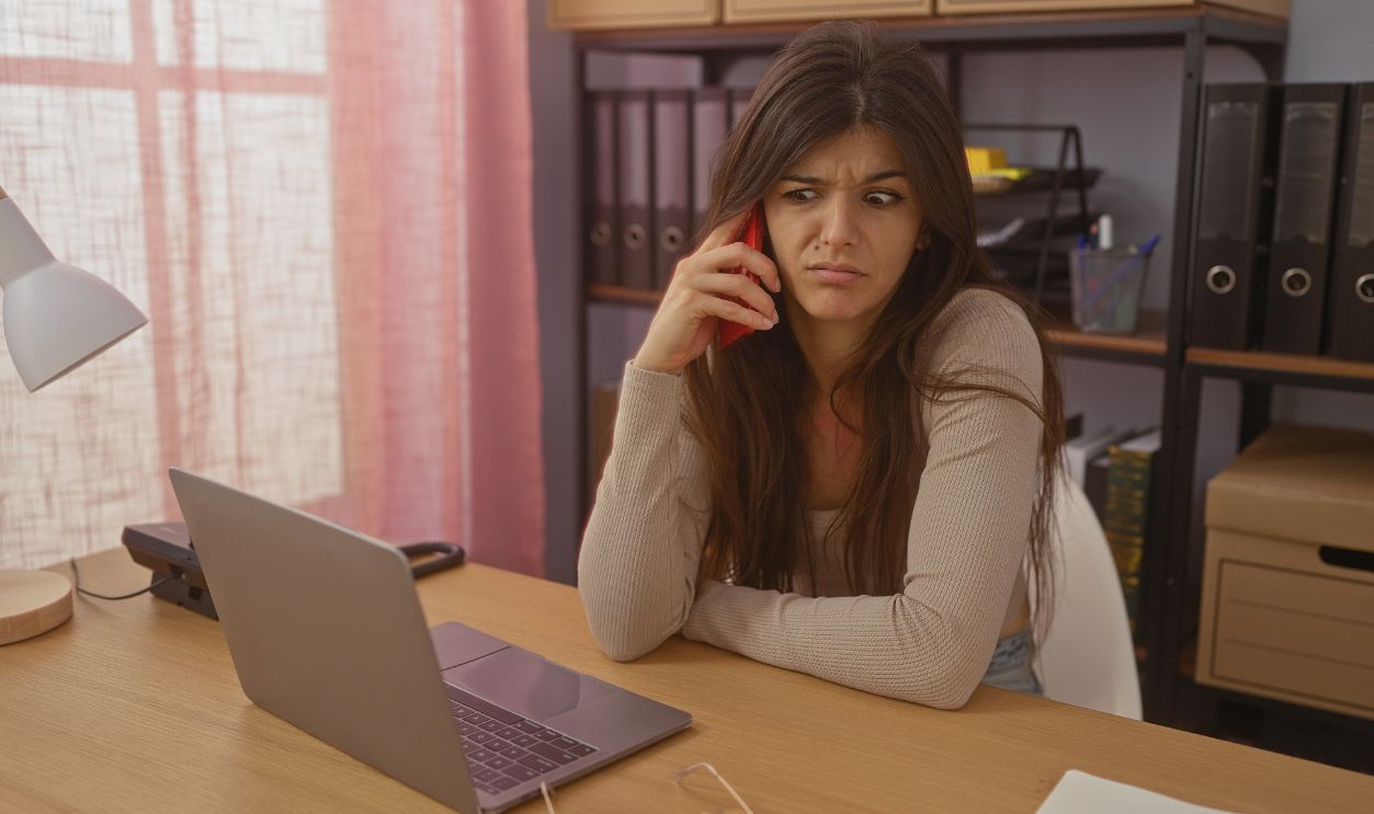 Young brunette woman at home office indoors looking concerned while talking on the phone with a laptop in front of her.