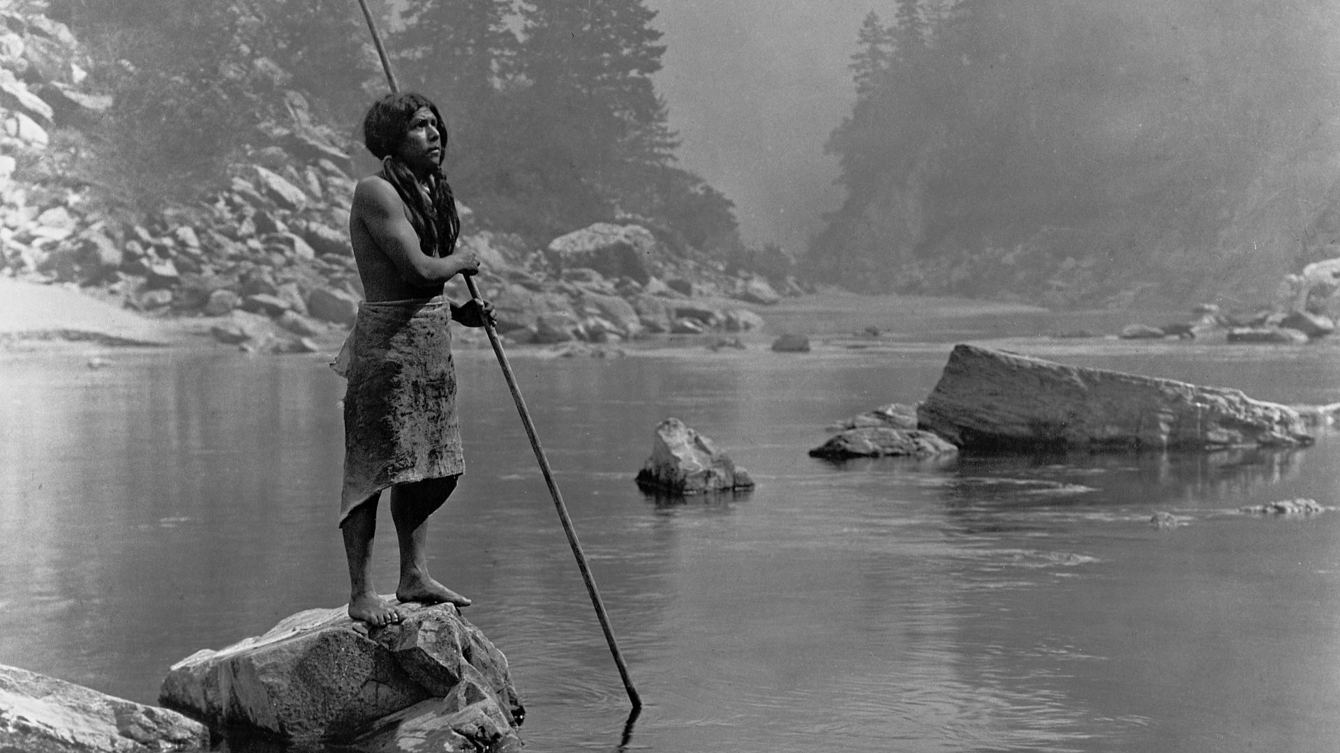 A smoky day at the Sugar Bowl--Hupa. Hupa man with spear, standing on rock midstream, in background, fog partially obscures trees on mountainsides. Published 1924 in The North American Indian / Edward S. Curtis. [Seattle, Wash.] : Edward S. Curtis, 1907-3