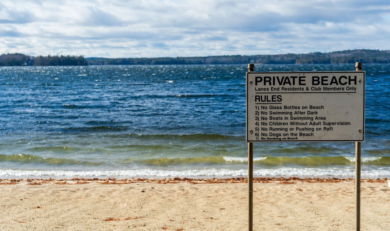 A private beach sign listing rules beside a scenic waterfront view under a cloudy sky.