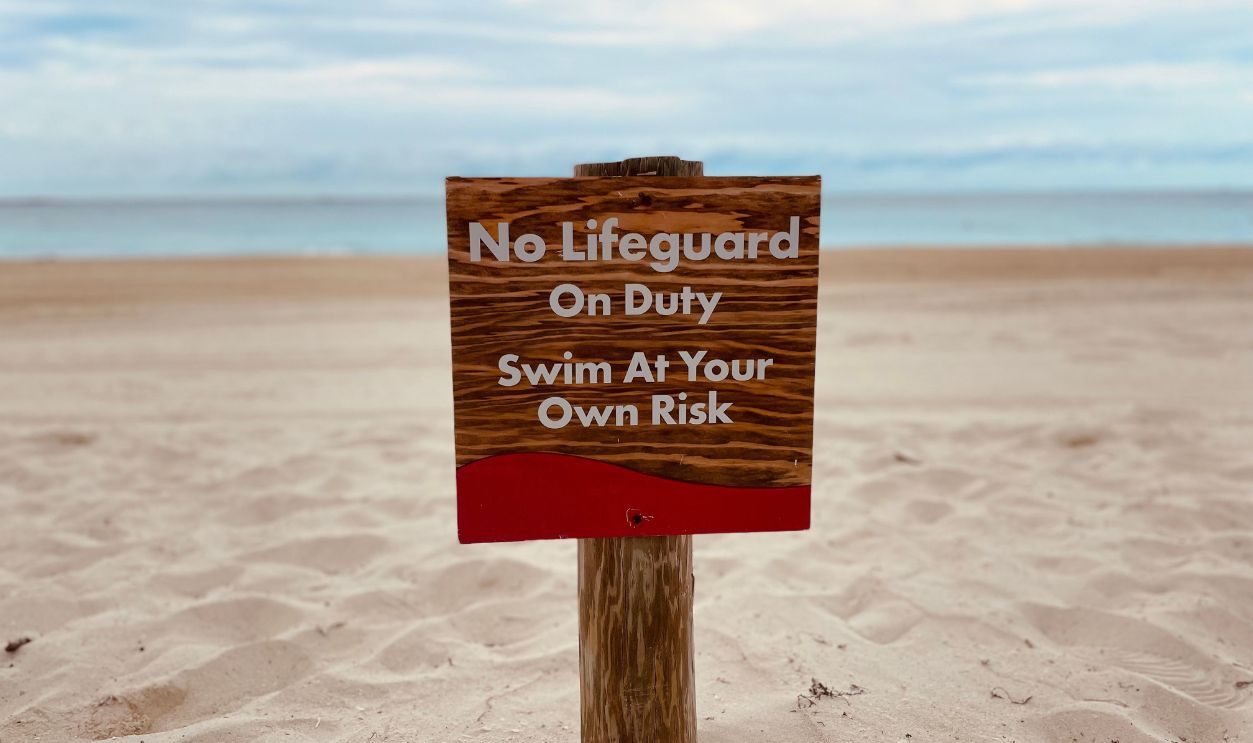 A sandy beach with a sign warning 'No Lifeguard On Duty - Swim At Your Own Risk.'