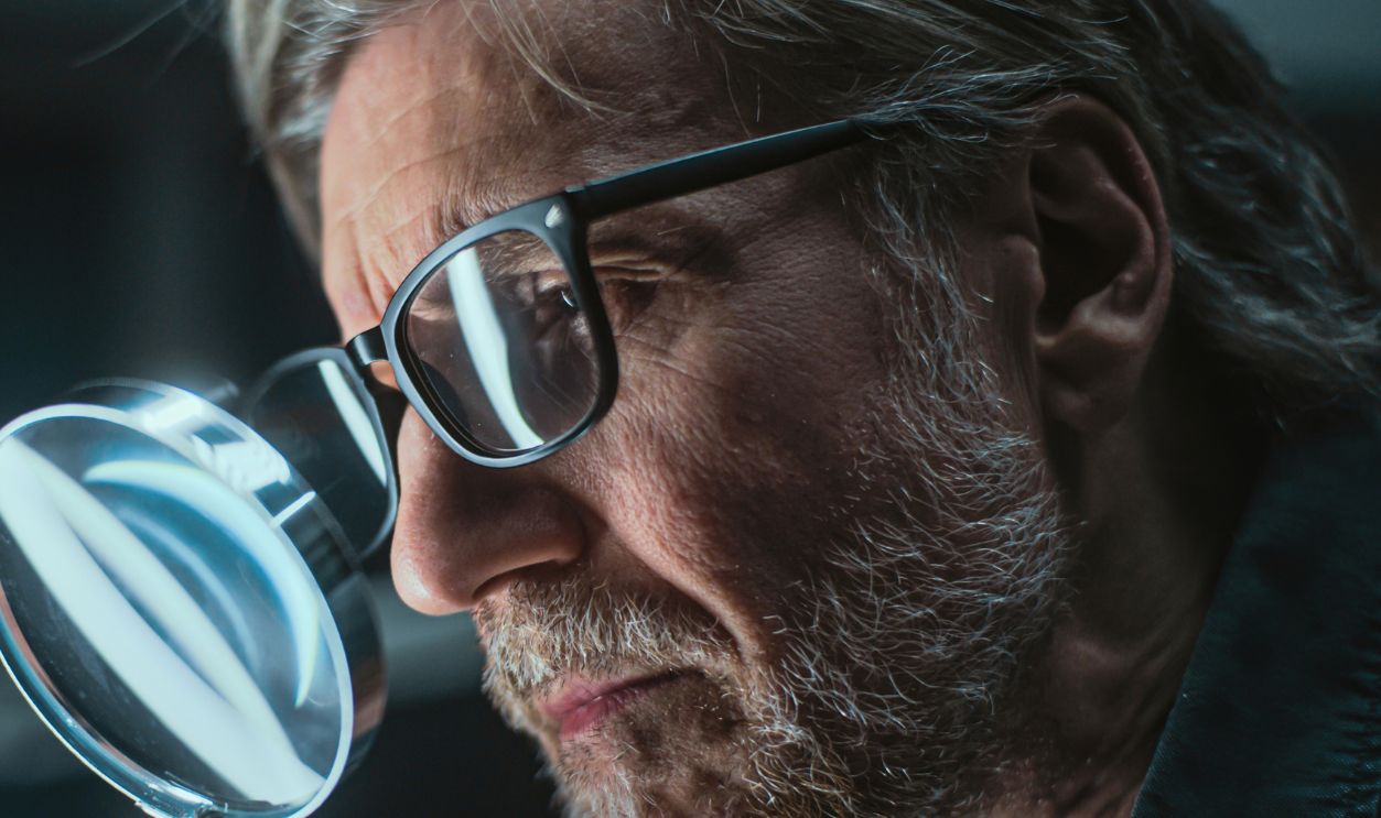 Mature archaeologist examines ancient extinct human or animal remains using magnifying glass. Scientist works with fossil, does scientific bone examination and research in archaeological lab at night.