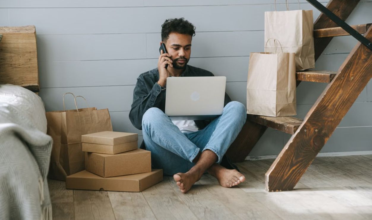 Adult man working remotely on laptop, surrounded by shopping bags and boxes indoors.