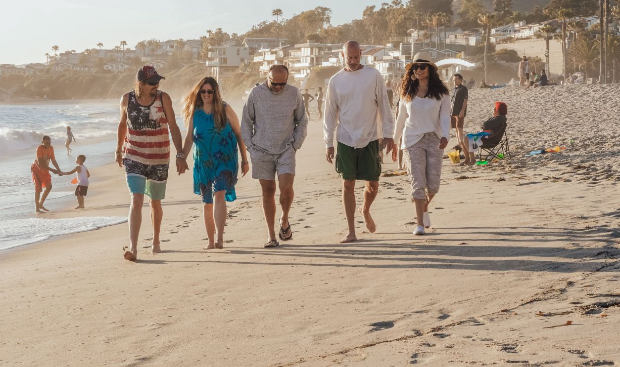 Five adults walk along a sunny beach, enjoying leisure time by the ocean in summer.