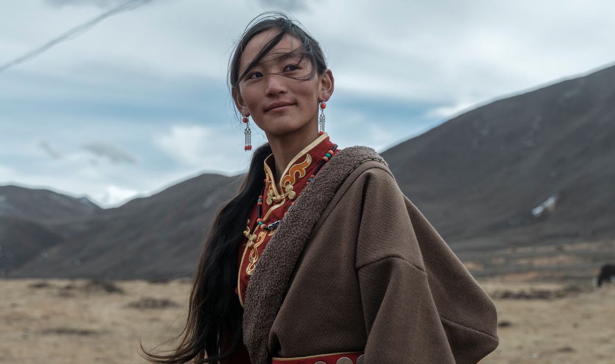 A young woman stands in a Tibetan landscape wearing traditional attire with decorative details