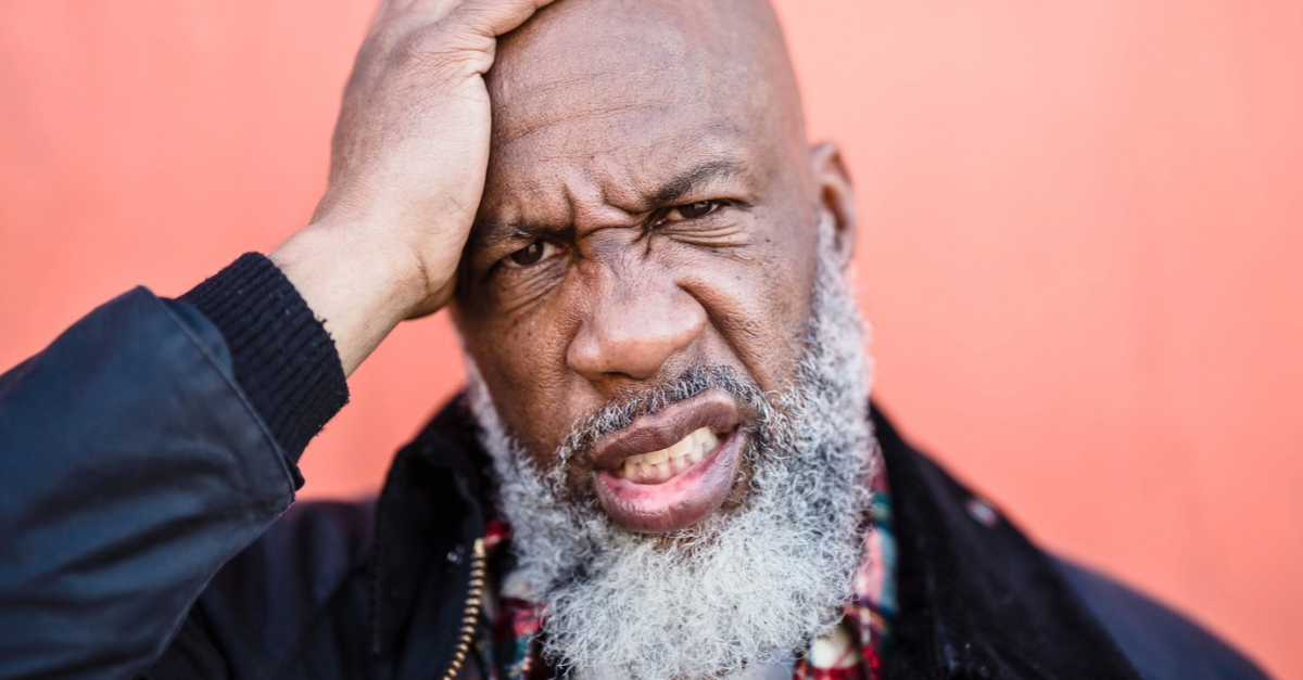 Close-up portrait of a bald, bearded man with a hand on his head, expressing emotion against a peach background.