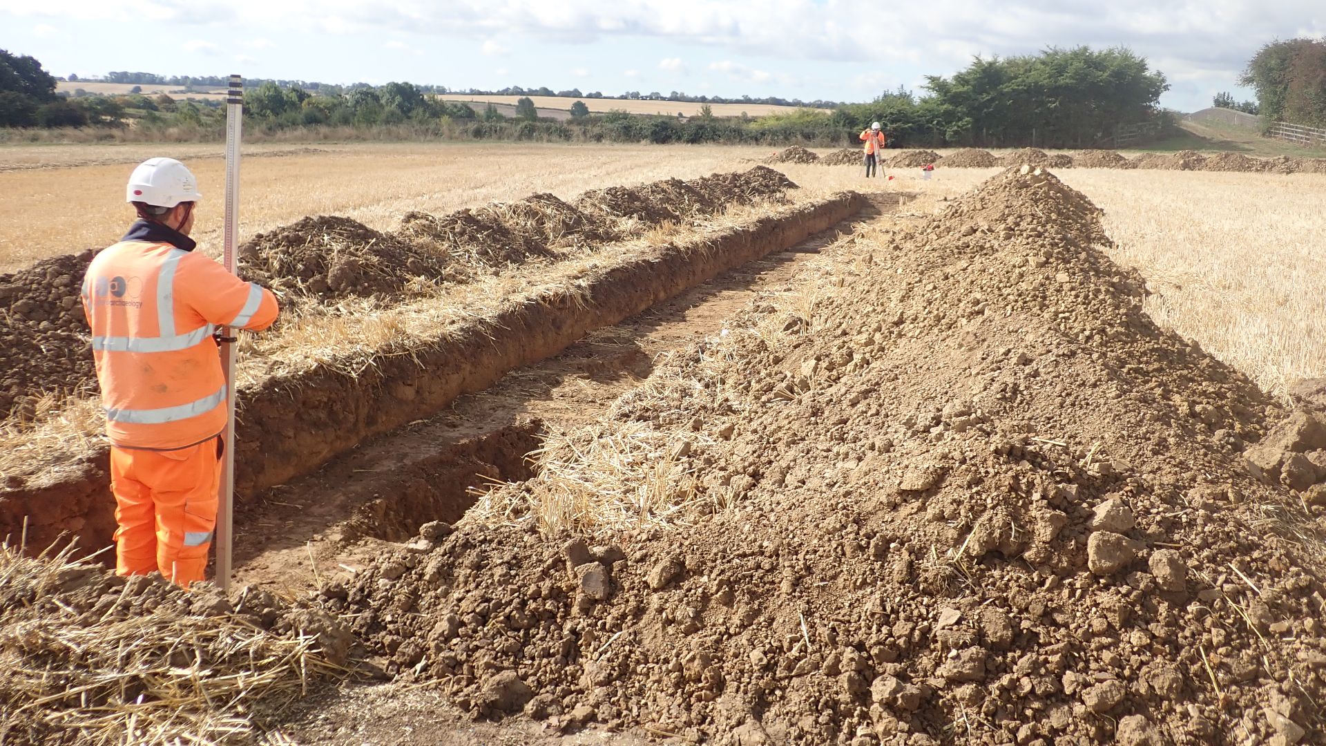 An archaeologist standing in the end of trench, holding a ranging rod as an archaeologist in the distance uses survey equipment.