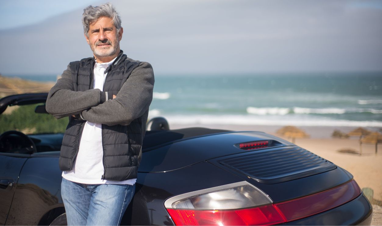 An elderly man stands confidently by his car on a scenic beach in Portugal.