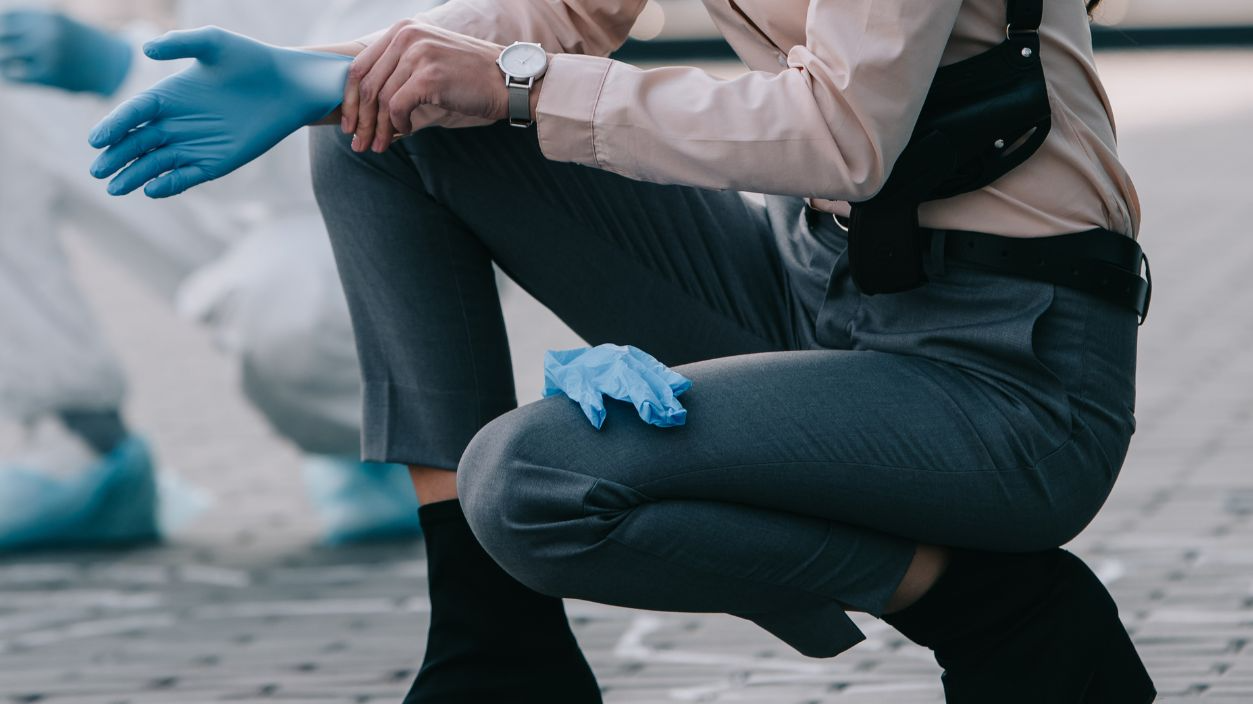 Shutterstock-1228738567, female detective in sunglasses wearing latex gloves with criminologist behind at crime scene