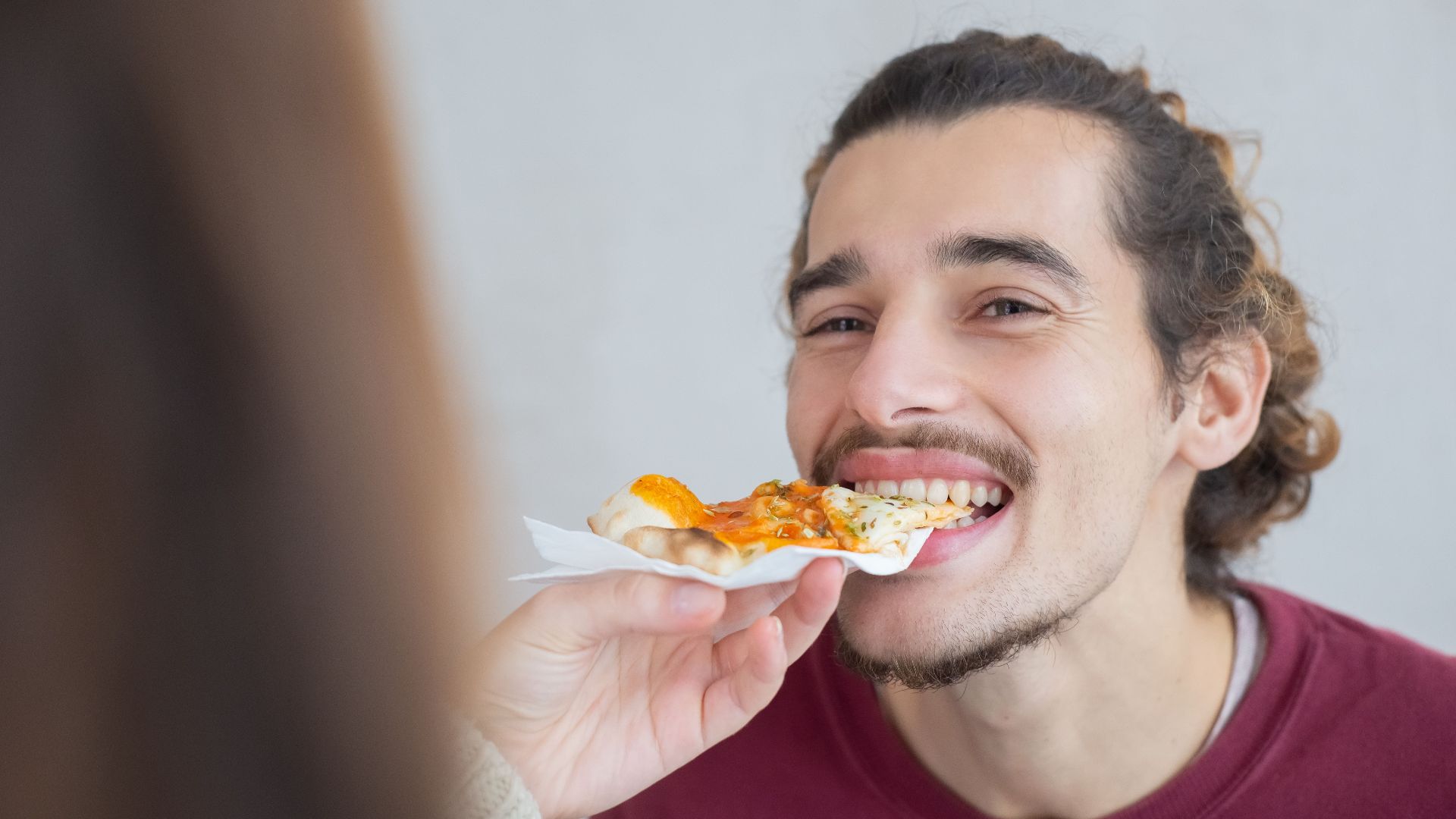 Smiling young man with facial hair enjoying a slice of pizza indoors.
