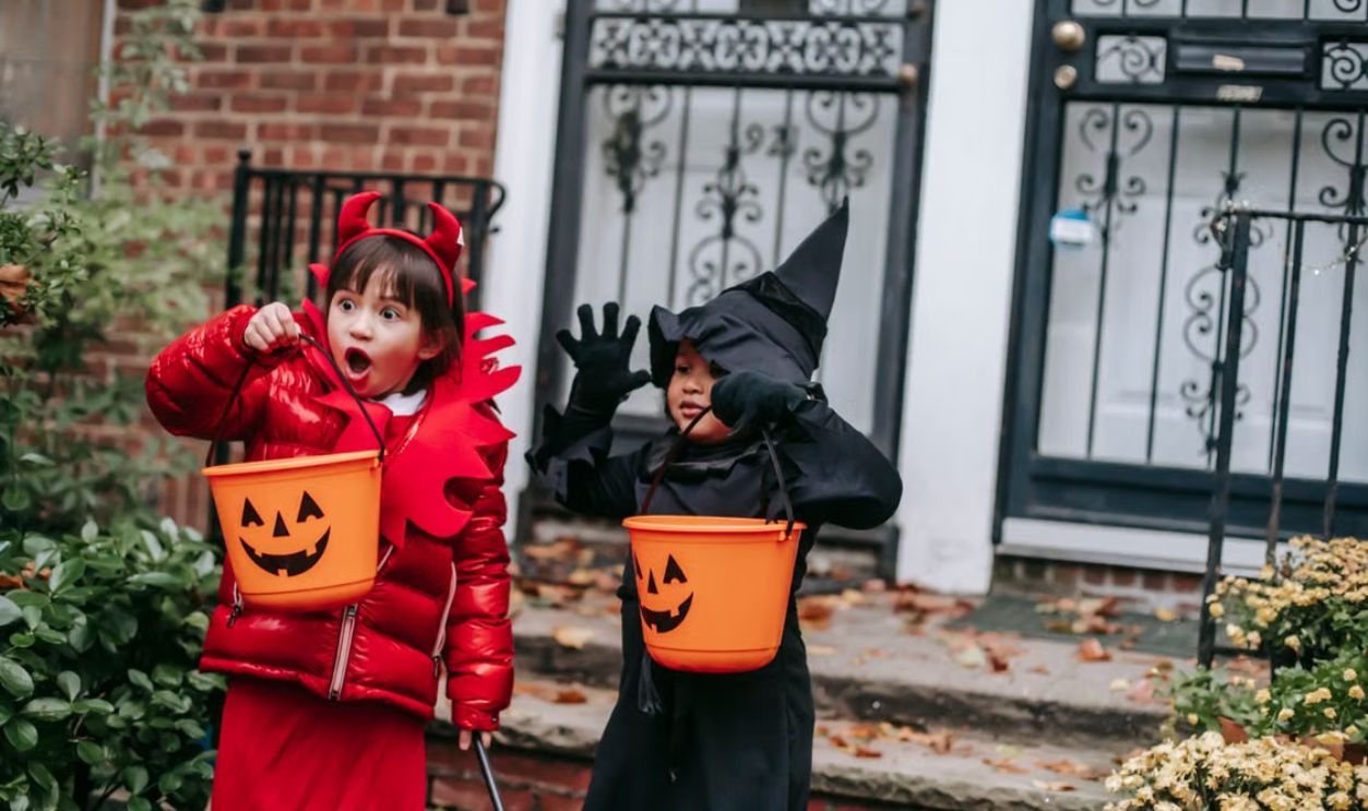 Two children in Halloween costumes trick-or-treating with pumpkin buckets outside a decorated house