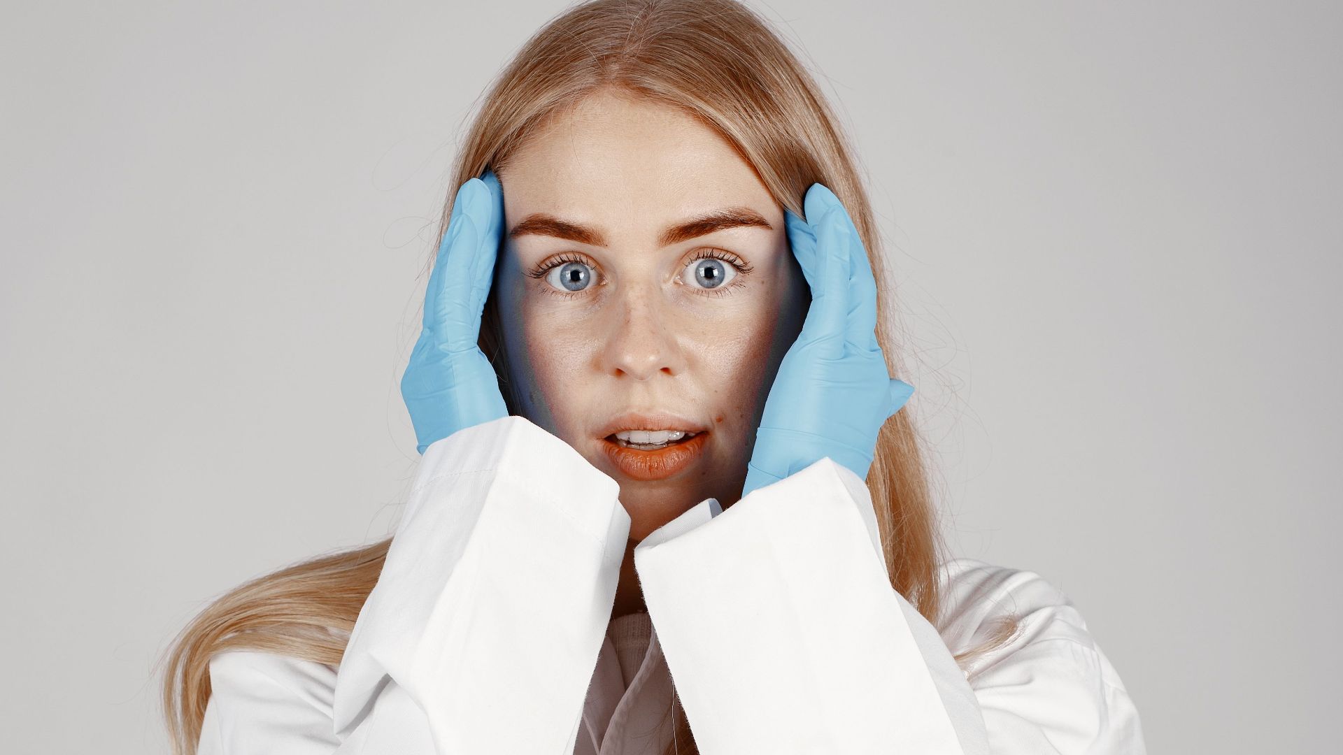 Young female scientist in white lab coat with blue gloves looking surprised.