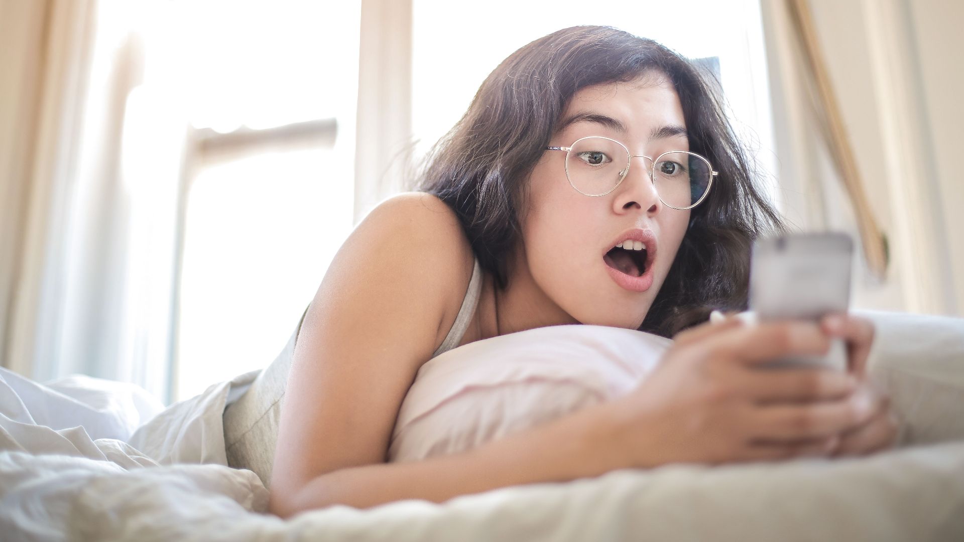 Young woman lying on bed, surprised by something on her smartphone, in a bright bedroom setting.