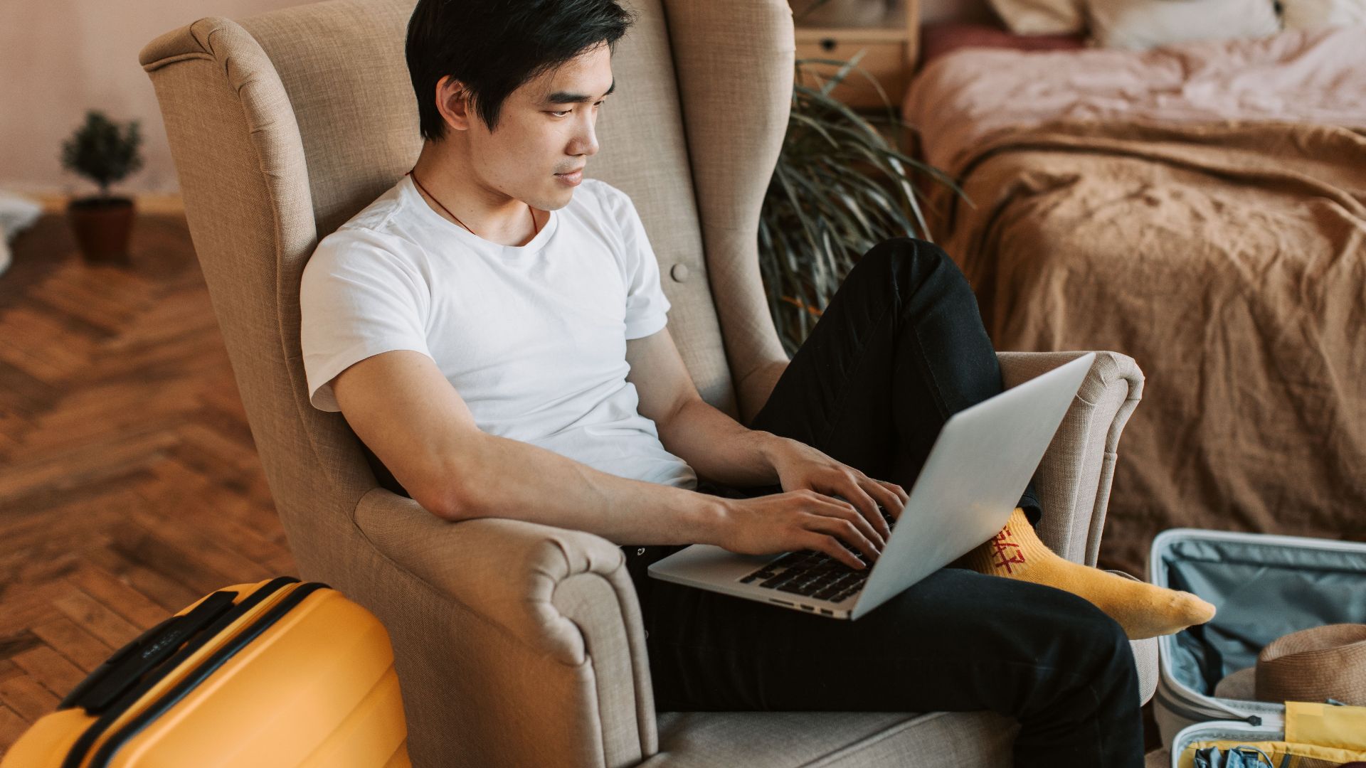 A man sitting comfortably in an armchair, using a laptop in a cozy home interior.