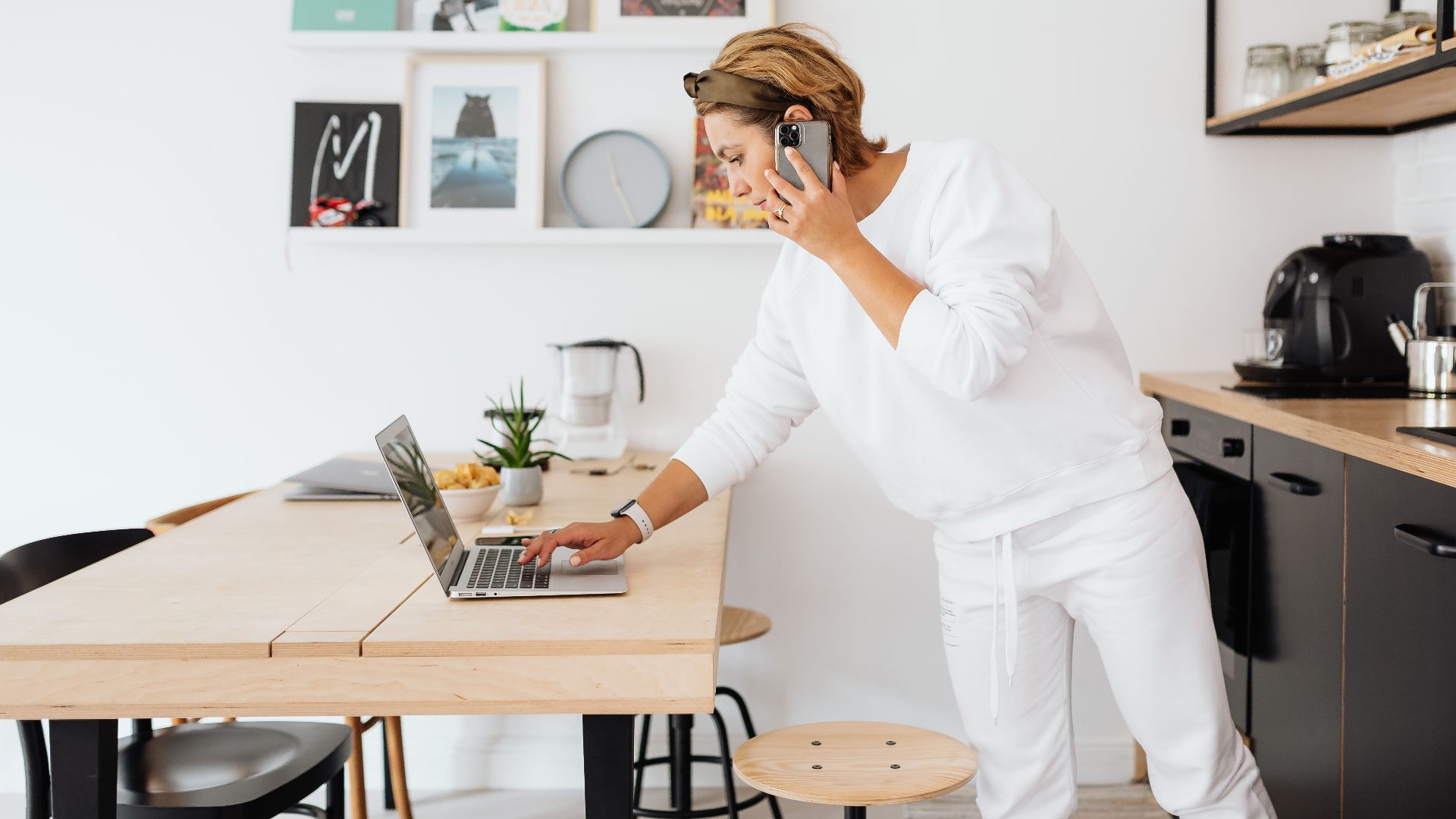 Woman using laptop and phone in stylish kitchen, blending work and home life.