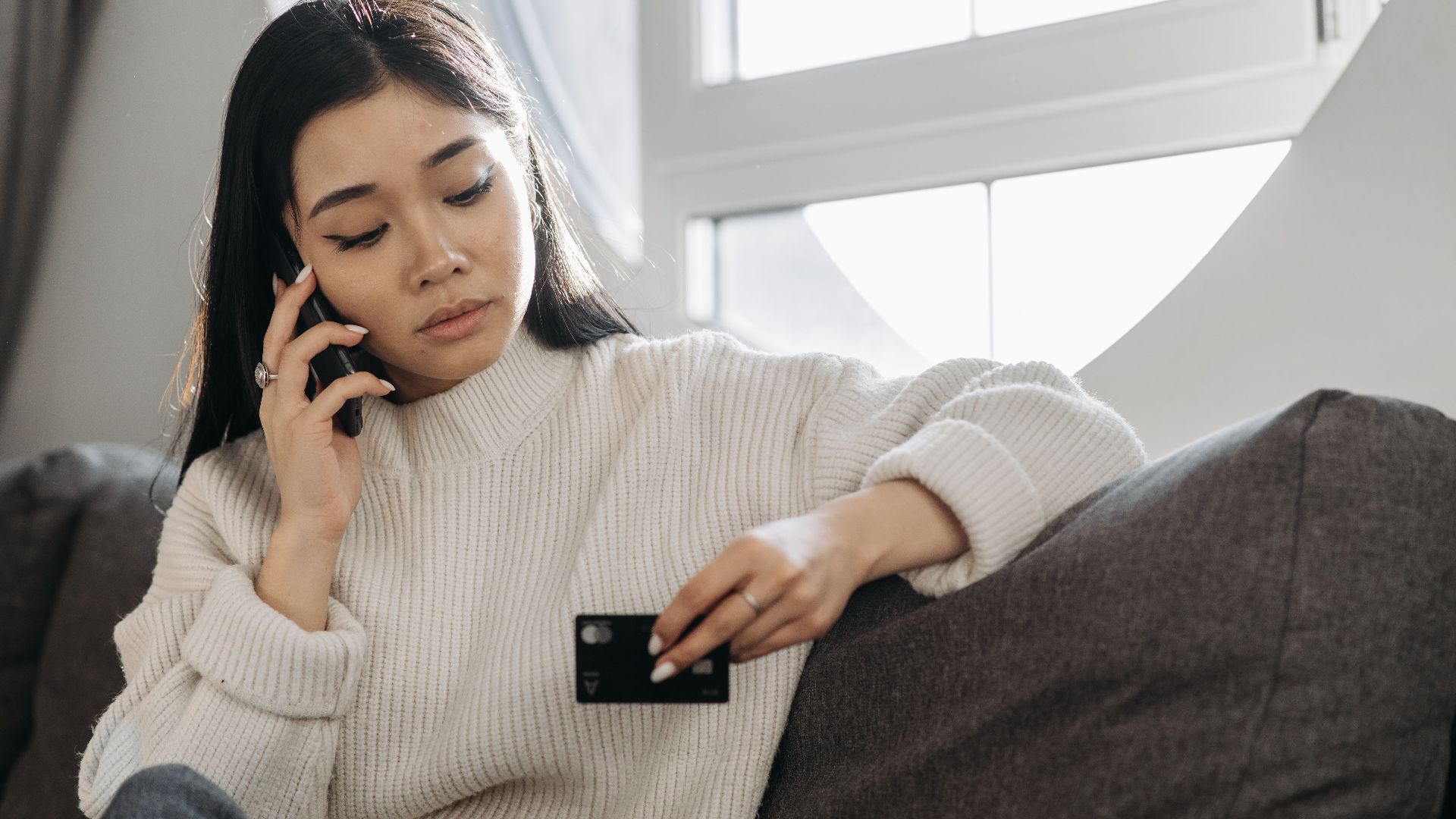 A woman sits on a sofa, multitasking with a phone and credit card, focusing on online shopping.