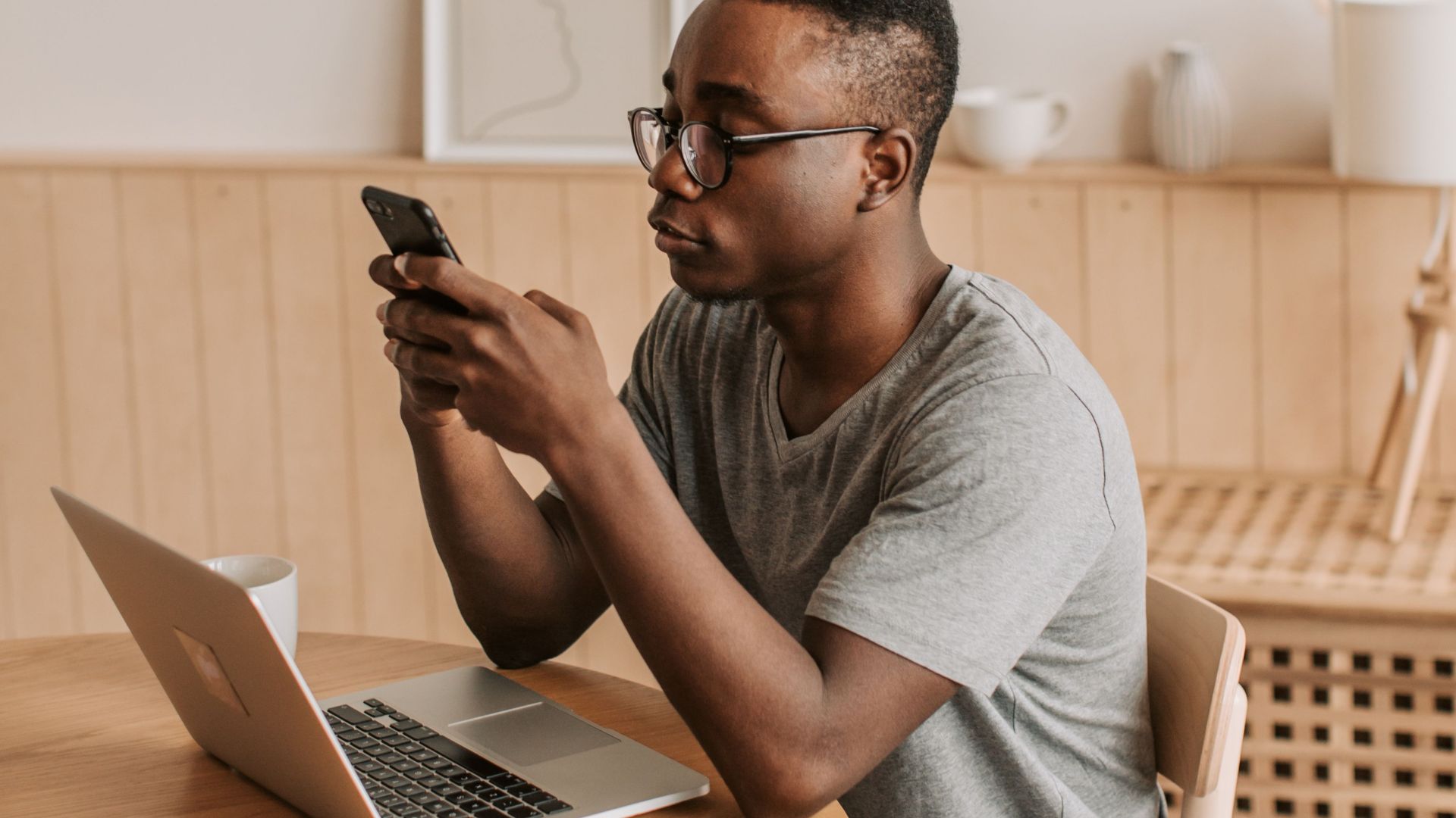 Young man in glasses using smartphone indoors, working on laptop at a modern wooden table.