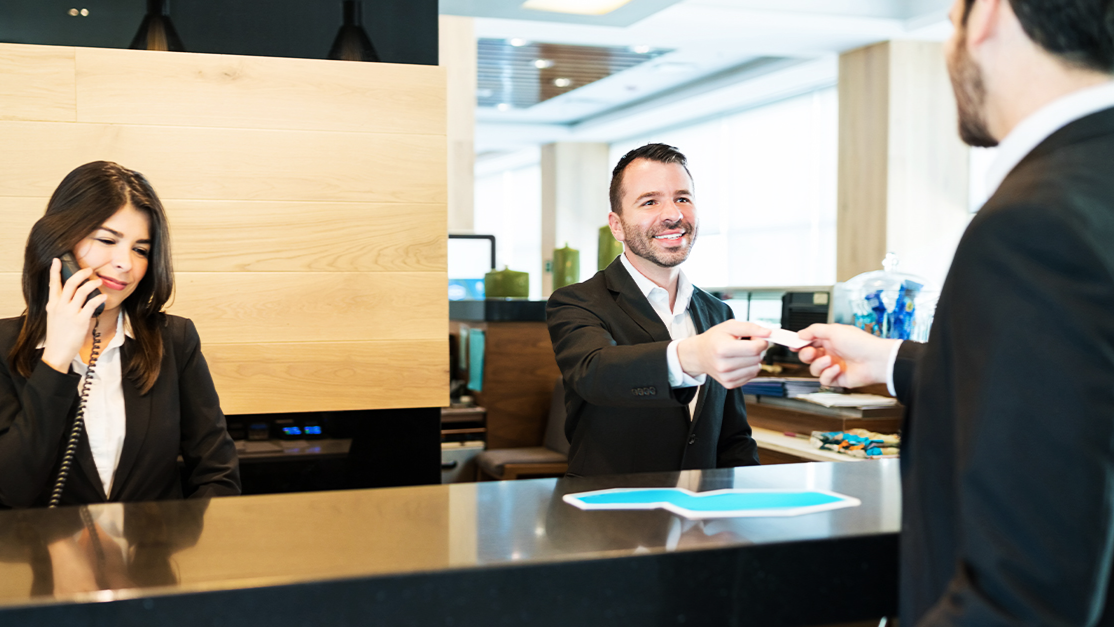 Smiling hotel front desk agent giving access key to guest checking in, while colleague talks on phone at hotel reception.