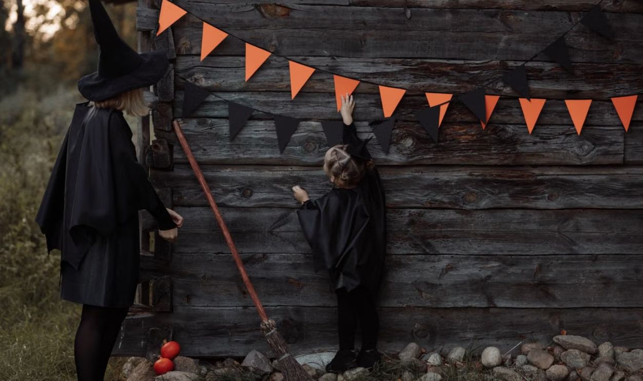 A mother and daughter in witch costumes hang pennant banners on a wooden wall for Halloween