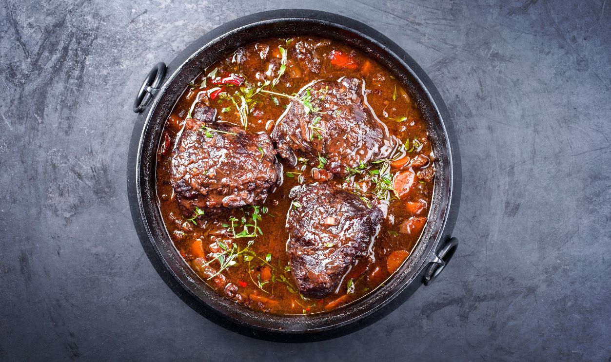 Traditional German braised beef cheeks in brown red wine sauce with carrots and onion offered as top view in a modern design stewpot on an old rustic board