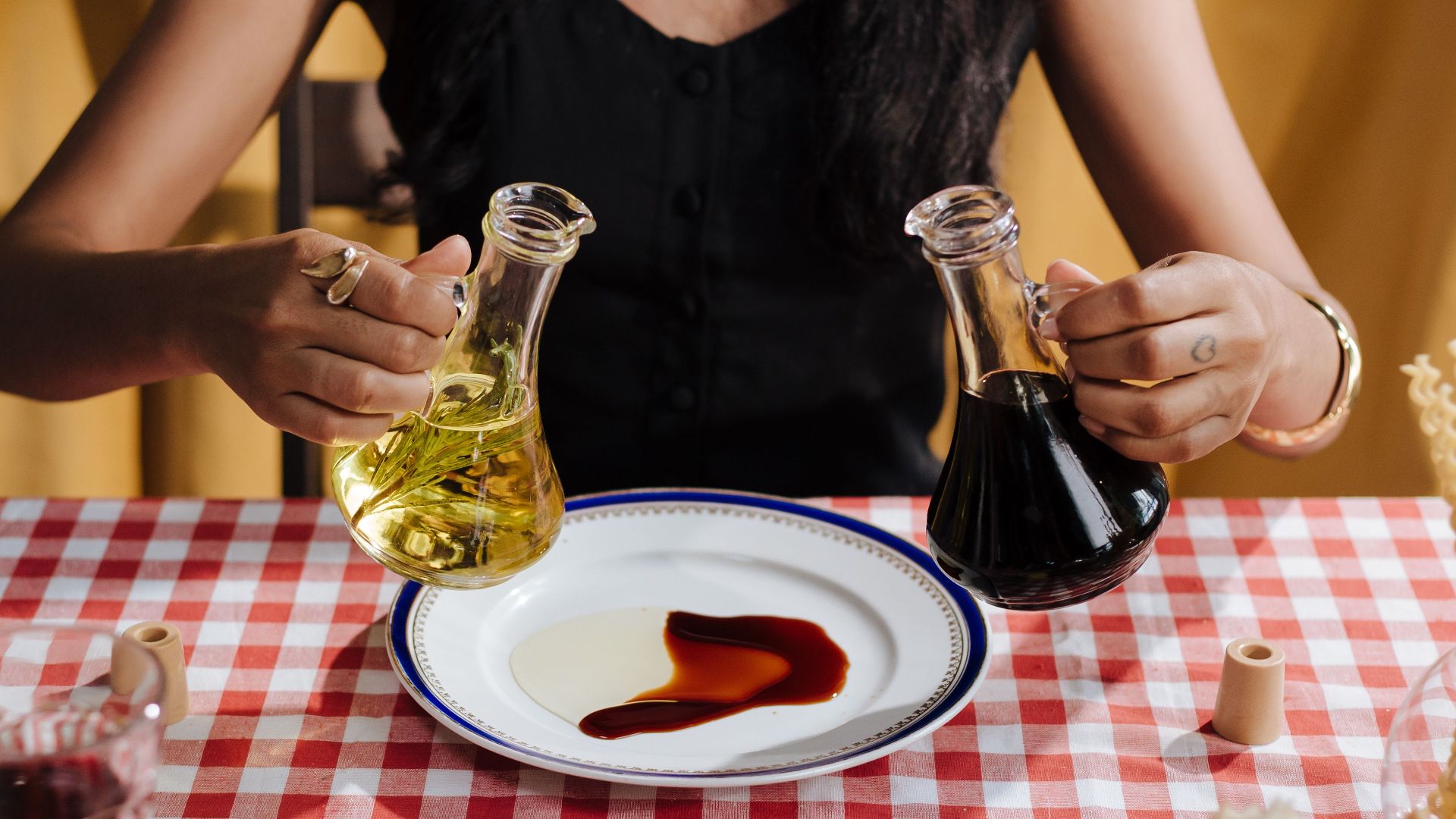 A woman pours olive oil and vinegar on a plate over a red checkered tablecloth.